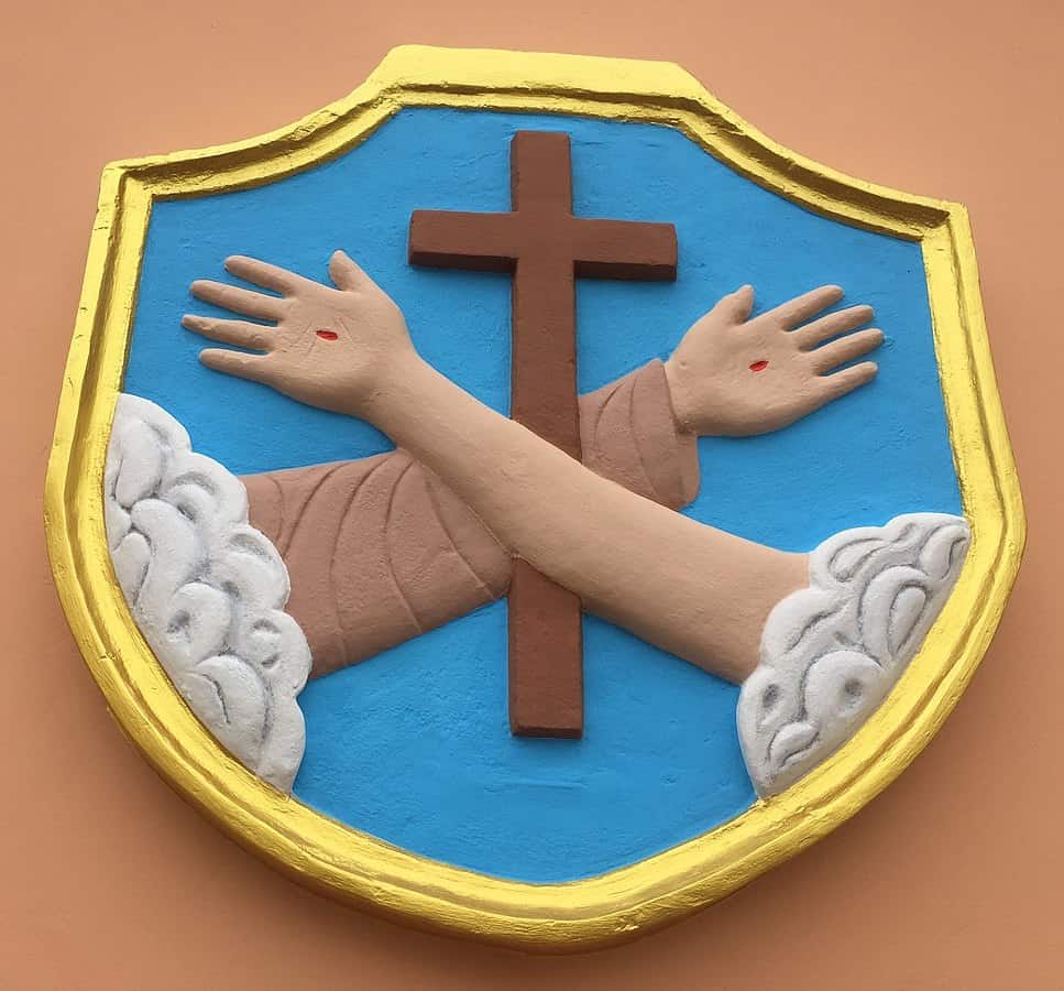 The image displays a relief sculpture of the Franciscan Coat of Arms from Mater Dolorosa Church. This emblem features a blue shield with a central brown cross, flanked by two arms—one clothed in a Franciscan robe and the other bare—both showing wounds on the hands, symbolizing St. Francis of Assisi's stigmata. Above and below the arms are white clouds. The shield is edged with a golden border, enhancing its distinct and symbolic design. This artistic representation is rich in religious symbolism, reflecting the Franciscan commitment to humility, peace, and service.